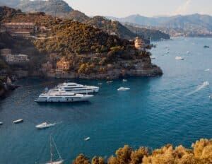 A stunning scene typical of a luxurious yacht charter in Corsica. The picture shows multiple yachts anchored near a rugged coastline adorned with lush vegetation and terracotta-roofed buildings. The water is a vibrant shade of blue, reflecting the unspoiled beauty of the Mediterranean landscape.